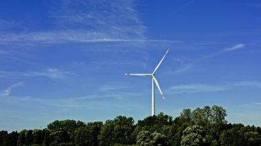 Uzun bir rüzgar türbini, berrak mavi gökyüzünün altında canlı yeşil ağaçların çerçevelediği hafif meltemde yavaşça döner. Huzurlu sahne doğanın ahengi ve yenilenebilir enerjiyi yakalar.