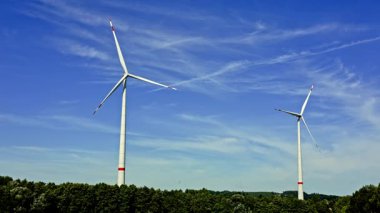 Yükselen iki rüzgar türbini, rüzgarda yavaşça dönen pervaneleri, parlak mavi gökyüzü ve çevreleyen yeşil ağaçlar yenilenebilir enerjiyi ön plana çıkaran barışçıl bir atmosfer yaratıyor.