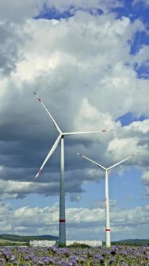 Büyük rüzgâr türbinleri, yeşil bir alanda büyük kabarık bulutlarla dolu parlak bir gökyüzünün altında nazikçe döner.