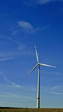 Geniş bir arazinin üzerinde, yumuşak bulutlu, mavi bir gökyüzünün altında uzun bir rüzgar türbini kuleleri. Sahne, sakin bir kırsal alanda yenilenebilir enerjinin özünü yakalıyor.