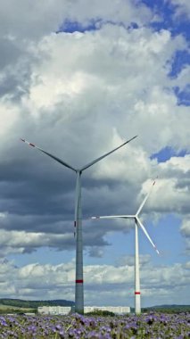 İki rüzgar türbini belirgin bir şekilde yükseliyor. Rüzgârda yavaşça dönen pervaneleri, dingin bir ortamda yenilenebilir enerjiyi ön plana çıkaran yumuşak bulutlara çarpıyor.