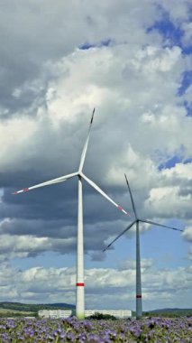 İki uzun rüzgar türbini renkli çiçeklerle dolu verimli bir arazide dönerken görülür gökyüzü büyük yumuşak bulutlarla doludur barışçıl bir atmosfer yaratır.