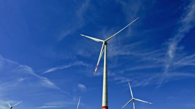 Uzun rüzgâr türbinleri, meltemde yavaşça dönerek yükselirler. İnce bulutlarla dolu berrak mavi gökyüzüne karşı yenilenebilir enerjinin gücünü gösterirler.