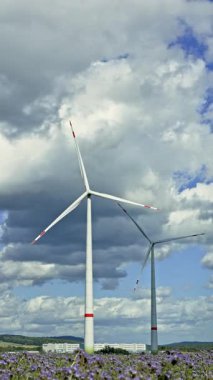 İki büyük rüzgar türbini, yumuşacık bulutlardan oluşan bir zemin ve çiçek açan bir çiçek tarlasına karşı zarifçe dönerek doğayla uyum içinde yenilenebilir enerjinin huzurlu bir sahnesini oluşturuyor.