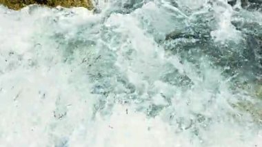 Parlak güneş ışığı, dalgalı okyanus dalgalarından yansıyarak kıyı boyunca uzanan engebeli kayalara çarpar. Sahne, suyun ritmik sesiyle enerji ve sükunet taşır.