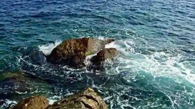 Kıyıdaki pürüzlü kayalara çarpan güçlü dalgaların yakından görüntüsü berrak bir gökyüzü ve güneş ışığıyla okyanusun canlı mavi rengini yükselten beyaz köpük ve dalgalanmalar yaratıyor.