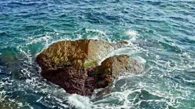 Sahil manzarasında berrak mavi dalgalar, güneş ışığını yansıtan engebeli bir kayanın üzerinde yuvarlanır. Huzurlu su, sonsuz bir dansla sahil şeridinde beyaz köpük oluşturur.