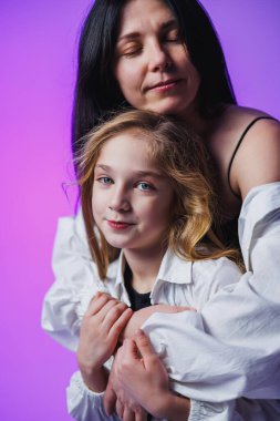 Sevgi dolu bir anne, kızına sıkıca sarılır. İkisi de rengarenk bir mekanda ışık saçan mor parıltıdan zevk alırken hafifçe gülümserler..