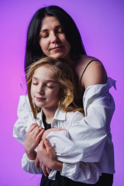 Bir anne ve kızı bir anı paylaşıyor, gözleri kapalı, sevgide kaybolmuşlar. Arkaplan yumuşak mor bir tonda parlıyor, bu samimi sahnede onların sevgi bağlarını güçlendiriyor..