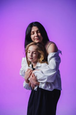 Bir anne, kızını yumuşak bir kucaklama ile kucaklar. Her ikisi de canlı mor bir arkaplan önünde huzur içinde gülümserler. Sevinç saçan huzurlu ve sevgi dolu bir ortam yaratır..