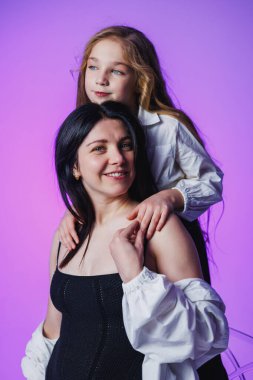 Bir anne ve kızı, annesinin omuzlarına tünemiş kızlarıyla neşeli bir anı paylaşıyor. İkisi de çarpıcı bir mor zemine gülümsüyor..