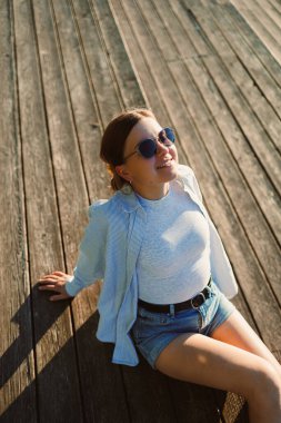 Neşeli bir kadın yıpranmış ahşap bir güvertede oturur, güneş ışığında sırılsıklam olur. Sade bir bluz ve şort giyerek, gamsız bir gülümsemeyle mutluluğu yayıyor, sıcak öğleden sonranın tadını çıkarıyor..