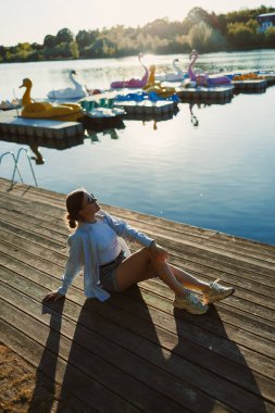 Genç bir kadın dingin bir gölün yanındaki ahşap iskelede güneşlenirken renkli kürek kayıkları yakınlarda yüzüyor ve huzurlu bir atmosfer yaratıyor..
