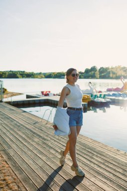 Genç bir kadın, sıcak güneşin altında göl kıyısında rahat bir yaz elbisesi giyerek geziniyor. Suyun kenarında eğlenceli bir güne hazır. Arka planda renkli tekneler var..