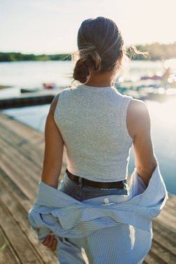 Genç bir kadın altın saatinde ahşap bir rıhtımda yürüyor, kolsuz bir bluz ve kot şort giyiyor, elinde mavi çizgili bir gömlek, göl manzarasının keyfini çıkarıyor..