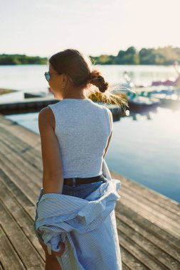 Genç bir kadın, huzurlu bir gölün kıyısındaki ahşap bir rıhtımda yürüyor, günlük yaz kıyafetleri giyiyor ve öğleden sonra güneşinin sıcacık ışığını kucaklıyor. Rahatlama havayı doldurur..
