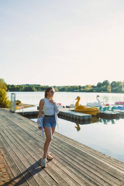 Bir kadın, sakin bir gölün kenarındaki ahşap rıhtımda yürüyor, sıcak güneş ışığının tadını çıkarıyor. Renkli kuğu şekilli tekneler yakınlarda yüzerek rahatlama ve doğa manzarası yaratıyor..