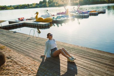 Genç bir kadın, güzel bir gölün kenarında, güneşin altında, ahşap bir rıhtımda rahatça oturur. Renkli kürek tekneleri yakınlarda süzülüyor, öğleden sonraki sakin atmosferi güçlendiriyor..