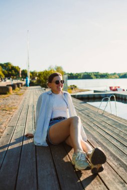 Genç bir kadın, gölün kenarındaki ahşap iskelede rahat bir şekilde oturur ve sıcak bir günde güneşin tadını çıkarır. Yaz kıyafetleri ve güneş gözlüğü takıyor. Kaygısız bir hava yayıyor..