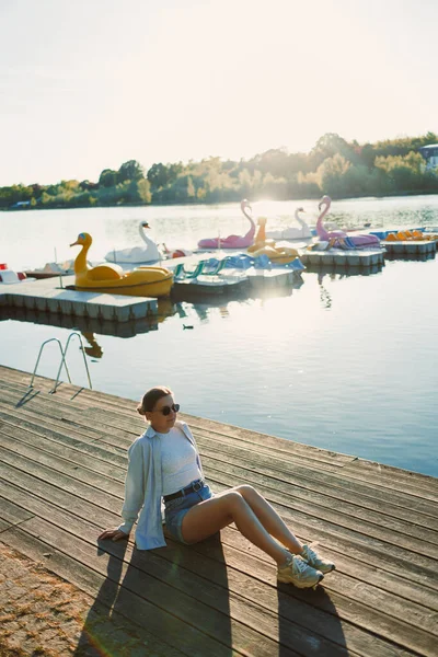 Bir kadın rıhtımda huzur içinde oturur, güneşi emer, canlı kürek tekneleri arka plandaki gölde usulca yüzer, sakin ve neşeli bir atmosfer yaratır..
