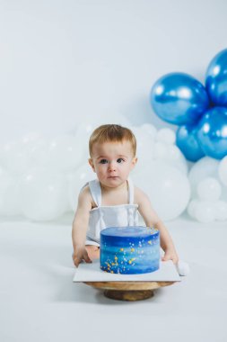 Tatlı bebek beyaz bir arka planda oturuyor, elinde güzel dekore edilmiş mavi bir pasta, altın aksanlı, neşeli mavi ve beyaz balonlarla çevrili..