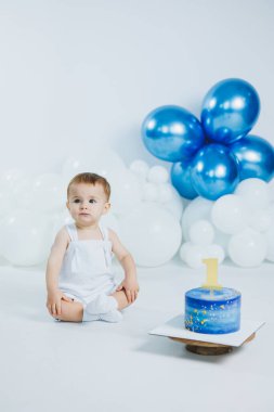 Bir yaşındaki neşeli bir çocuk yerde oturur ve üzerinde sarı bir mum bulunan güzel mavi doğum günü pastasına bakar. Beyaz ve mavi balonlar kutlamanın neşeli atmosferine eklenir.