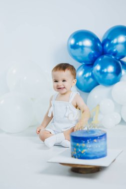 Bir yaşındaki neşeli bir çocuk, mavi ve beyaz balonlarla çevrili beyaz bir yüzeyde oturuyor. Bir numaralı rengarenk pastaya gülümseyip özel günlerini neşeyle kutluyorlar..