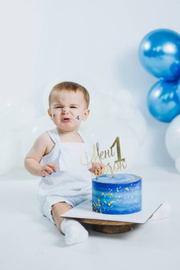 Mutlu bir bebek, mavi ve beyaz balonlarla çevrili parlak bir ortamda oturur. İlk doğum günlerini altın rengi bir pastayla kutlarlar..