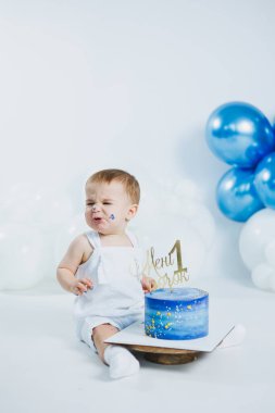 Neşeli bir bebek ilk doğum gününü mavi şekerli bir pasta, renkli balonlar ve yüzünde mutluluk ve şaşkınlık karışımıyla kutlar. Çocukluk merakının özünü yakalar..