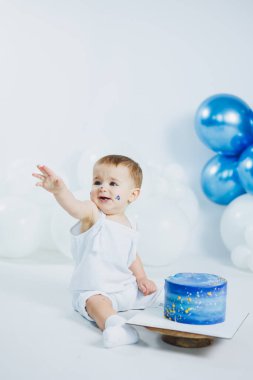 Beyaz elbiseli ufaklık, beyaz ve mavi balonlarla çevrili renkli mavi bir pastaya doğru mutlu bir şekilde el kol hareketi yapıyor..