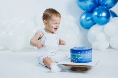 Genç bir çocuk, neşeli balonlarla çevrili güzel dekore edilmiş mavi bir pastaya uzanırken neşeli bir ifadeyle yerde oturuyor ve neşeli bir sahne yaratıyor..