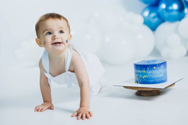 Neşeli bir bebek beyaz bir yüzey boyunca sürünüyor, yüzlerinde eğlenceli bir pasta şekerlemesi var. Yakınlarda, çarpıcı bir mavi pasta balonlarla süslenmiş şenlikli atmosfere canlılık katıyor..