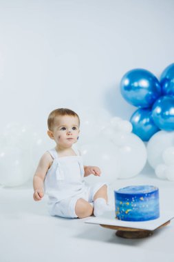 Beyaz tulumlu bir çocuk yerde oturmuş, balonlar arka planda süzülürken renkli süslemelerle süslenmiş mavi bir doğum günü pastasına bakıyor ve neşeli bir atmosfer yaratıyor..