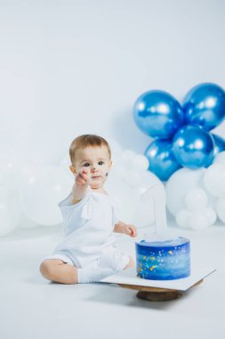 Beyaz elbiseli neşeli bir çocuk yerde oturuyor, mavi ve beyaz balonlarla çevrili güzel mavi bir pastayı işaret ediyor, ilk doğum günü kutlaması için..