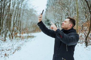 Sakin, karlı bir orman yolunda, neşeli bir suratı olan bir adam selfie çeker, çıplak ağaçlar ve yumuşak karlarla çevrili iken kışın büyüsünü yakalar..