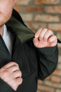 Genç bir adam sıcak tuğla bir duvarın önünde duruyor, parlak siyah ceketinin fermuarını dikkatlice kapatıyor. Doğal ışık odak noktasını ve kıyafetinin dokusunu vurgular..