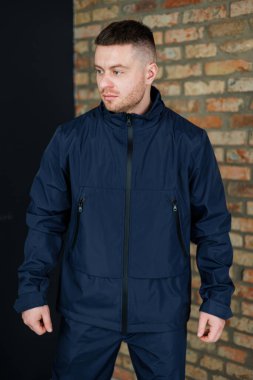 Koyu renk donanma ceketi giyen bir adam rahat ama modaya uygun bir görünüm sergiliyor. Sıcak tuğla duvar, şehir tarzını vurgulayarak modern sahneye doku ekler..