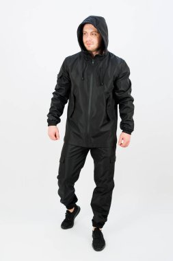 Parlak, siyah, su geçirmez bir ceket ve uyumlu bir pantolon giyen bir adam, parlak bir stüdyoda hazır bekliyor. Kıyafet, aktif yaşam tarzları için modern tasarım ve çok yönlülüğü vurguluyor.