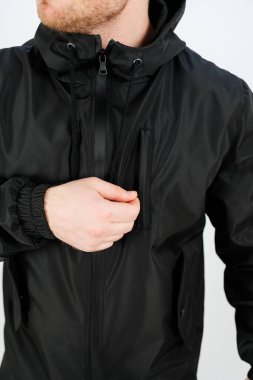 Bir kişi parlak siyah bir ceket giyer ve tarafsız bir arkaplanda dikilirken fermuarı tek eliyle düzeltir. Modern ve zahmetsiz görünüyor..