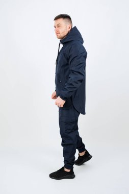 Genç bir adam rahat ama kendine güvenen bir duruşta duruyor. Çağdaş sokak modasını yansıtan parlak, koyu renk bir kıyafet giyiyor. İnce ışıklandırma sahneye şık bir hava katıyor..