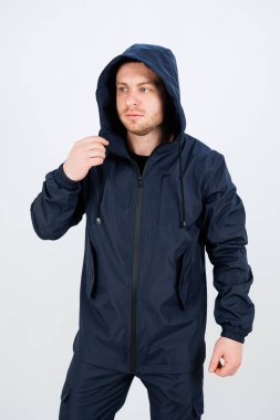 Donanma ceketi giyen bir adam, açık hava yolculuğuna hazırlanırken kaputu düzelterek kendinden emin bir şekilde duruyor. Basit bir arkaplan onun odaklanma ve kararlılığını vurguluyor..