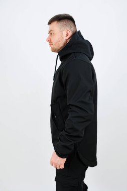 Genç bir adam gösterişli siyah bir kapüşonluyla duruyor, basit beyaz bir arkaplana karşı modern sokak kıyafetleri sergiliyor..