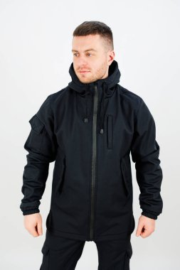 Kapüşonlu ve fermuarlı cepleri olan parlak siyah bir ceket giyen bir adam güvenle ayakta duruyor. İyi ışıklandırılmış stüdyo, onun modern tasarımını ve giyiminin uygunluğunu ön plana çıkarıyor..