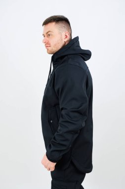 Tarz sahibi siyah kapüşonlu bir adam beyaz bir arka plana karşı poz verir. Kendine güveni modern moda eğilimlerini ve serin günler için ideal şehir tarzını vurguluyor..