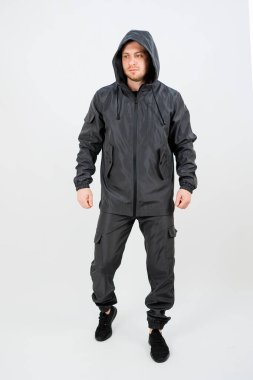 Model, kapüşonlu ve kargo pantolonlu şık, siyah atletik giysiler içinde duruyor. Basit bir ortamda konfor ve modern modaya vurgu yapıyor..