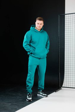 Kendine güvenen bir model şık bir turkuaz eşofman içinde parlak siyah beyaz bir arka plana karşı duruyor, konfor ve kentsel tarzın bir karışımını somutlaştırıyor. Rahat duruşu, çağdaş atmosfere yeni bir hava katıyor..