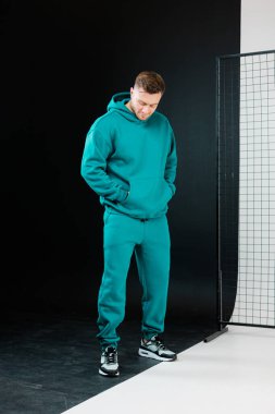 Bir model, parlak bir stüdyo arkaplanı ile çevriliyken, güvenle bir deniz mavisi eşofmanının içinde duruyor. Görünüm, çağdaş sokak giyimi eğilimlerini vurguluyor.