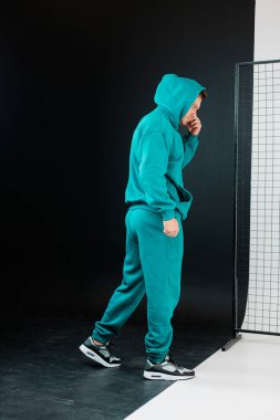 Deniz mavisi eşofman giyen bir adam düşüncelere dalmış, derin düşüncelere dalmış, zıt siyah bir arkaplanı olan canlı rengi yükselten bir adam. Sıradan görünüşü çağdaş bir moda havası yakalıyor..