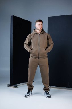 Şık bir adam, elleri cepte, şık arka planlarla çevrili, günlük kıyafetlerle gururla durur. Rahat duruşu çağdaş bir stüdyo ortamında kendine güvenini gösteriyor..