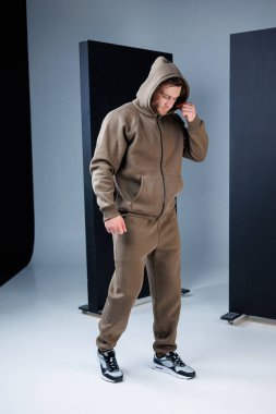 Genç bir adam modern bir stüdyoda kapüşonlu kahverengi eşofmanını sergiliyor. Kıyafetini ayarlıyor, rahat ama şık bir hava yayıyor..